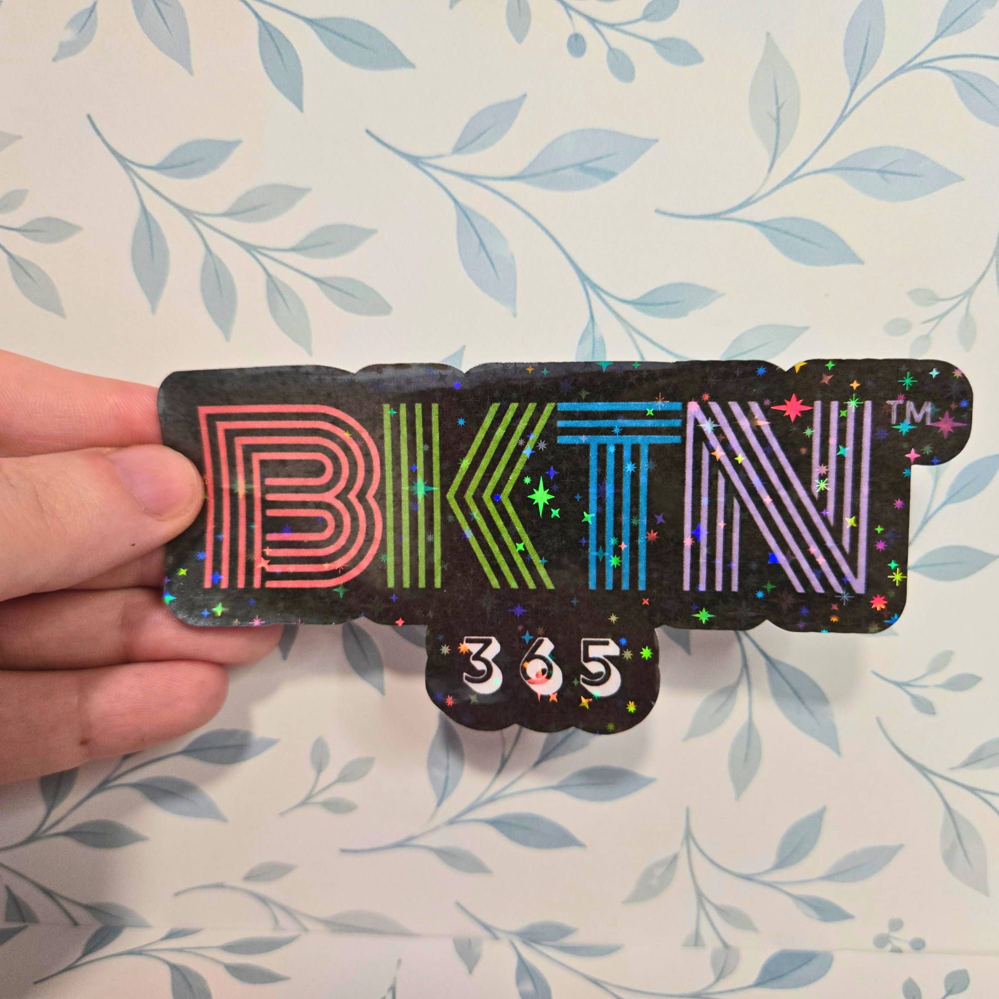 Sticker with 'BKTN' and '365' text held by a hand against a floral background