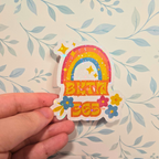 Colorful sticker with rainbow, stars, and flowers held by a hand against a floral background