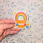 Hand holding a colorful sticker with a rainbow and text against a floral-patterned background
