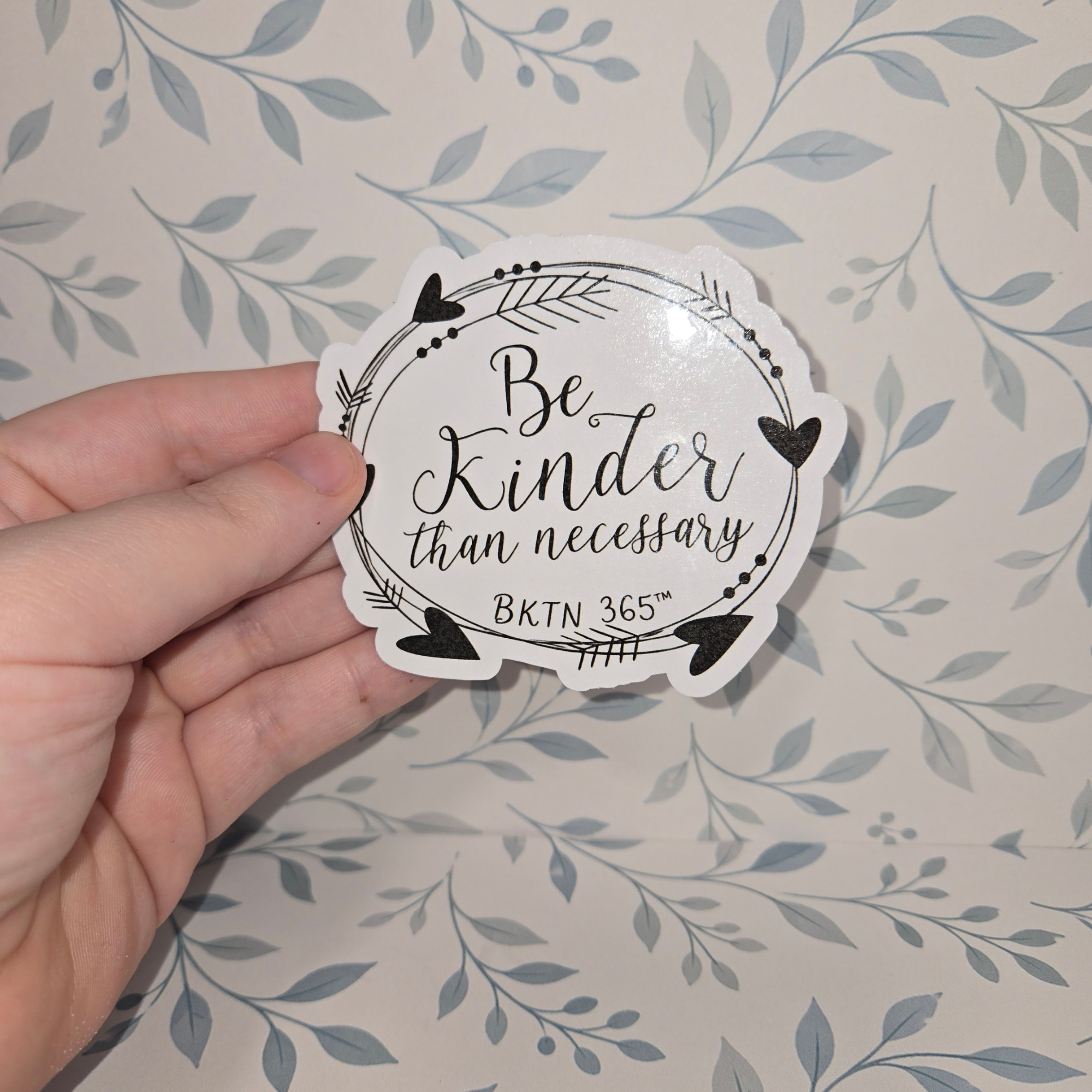 Hand holding a sticker with 'Be Kinder than necessary' text against a leaf-patterned background