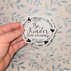 Hand holding a circular sticker with 'Be Kinder than necessary' text against a floral background
