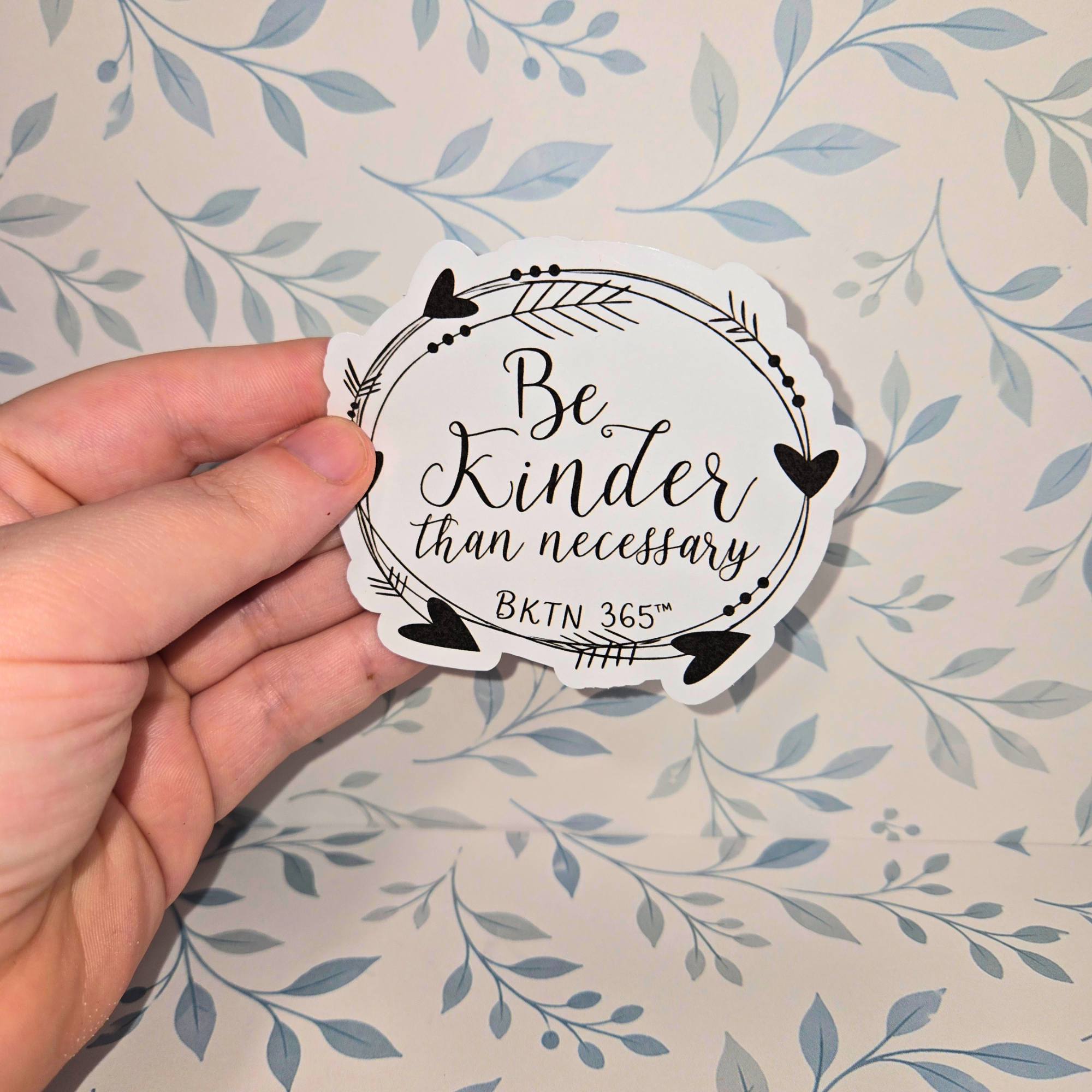 Hand holding a circular sticker with 'Be Kinder than necessary' text against a floral background